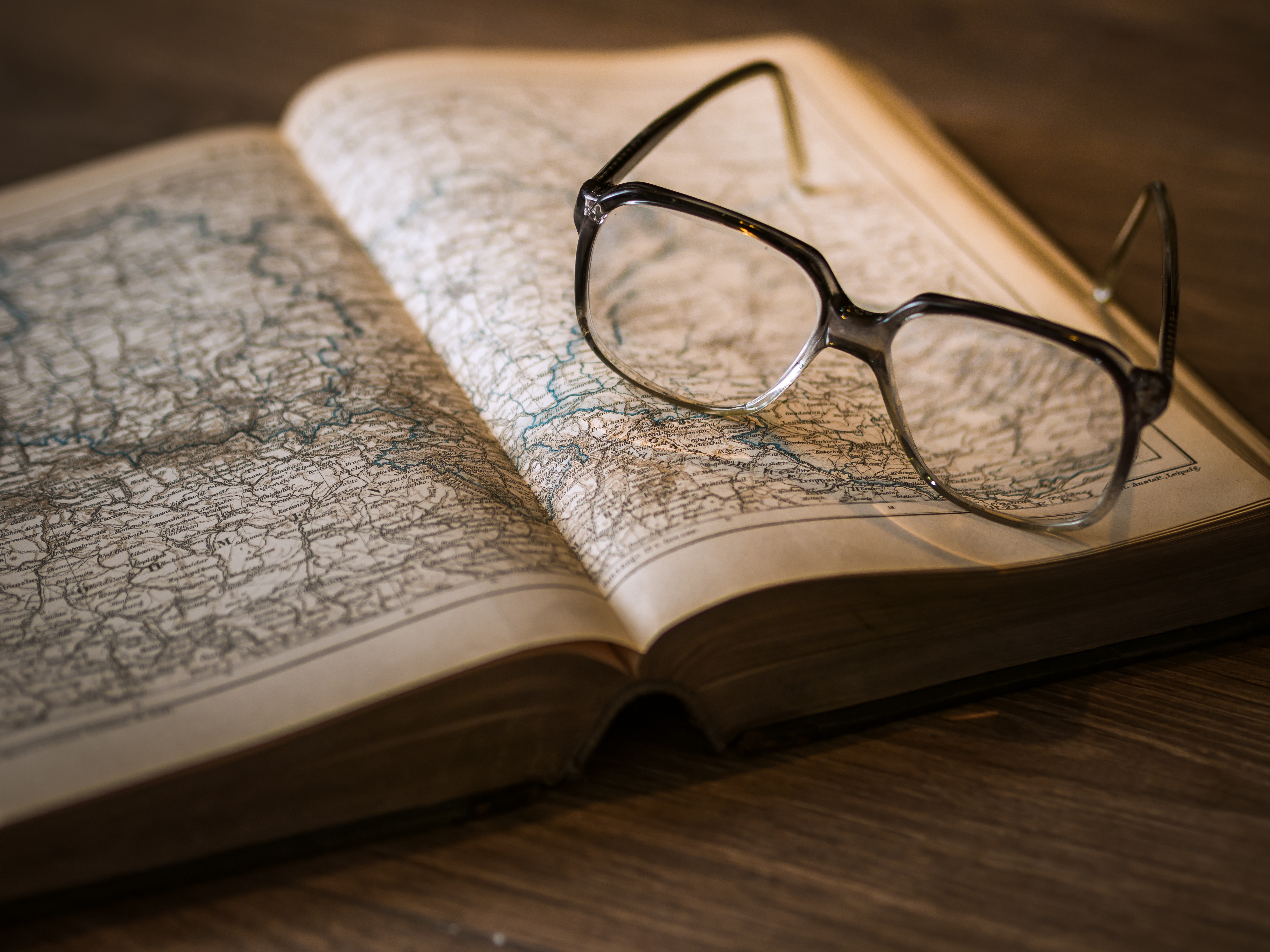 Une paire de lunettes posée sur un livre ancien ouvert, montrant des cartes détaillées, sur une table en bois sous une lumière chaleureuse.