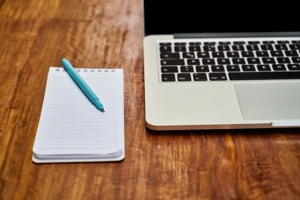 Un ordinateur portable ouvert à côté d'un petit carnet de notes blanc et d'un stylo bleu turquoise sur un bureau en bois rustique.