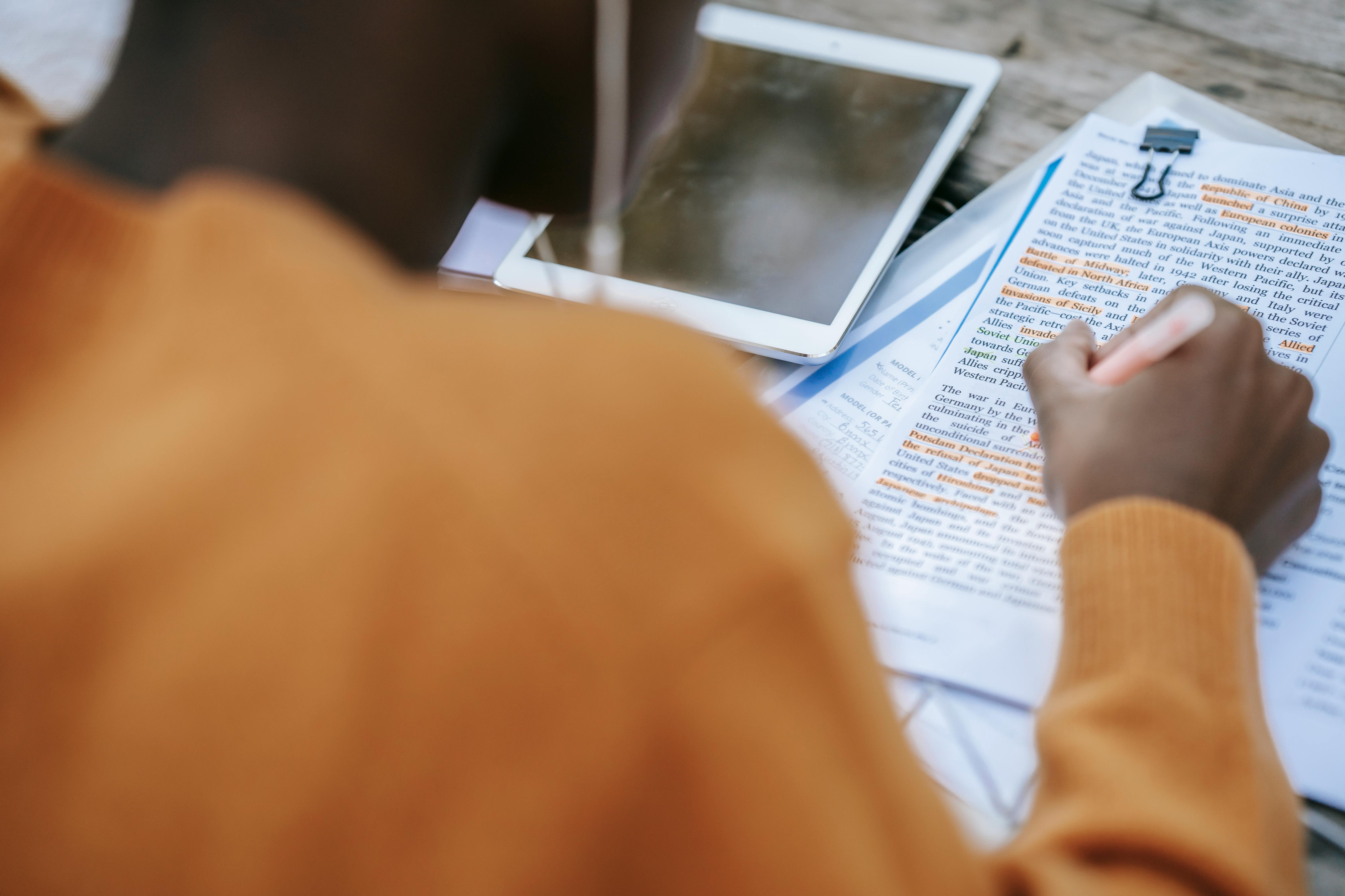 Vue de dessus d'une personne de dos, vêtue d'un pull moutarde, en train d'étudier. Elle utilise un surligneur rose sur un document papier rempli de texte sur la Seconde Guerre mondiale, à côté d'une tablette numérique posée sur la table.