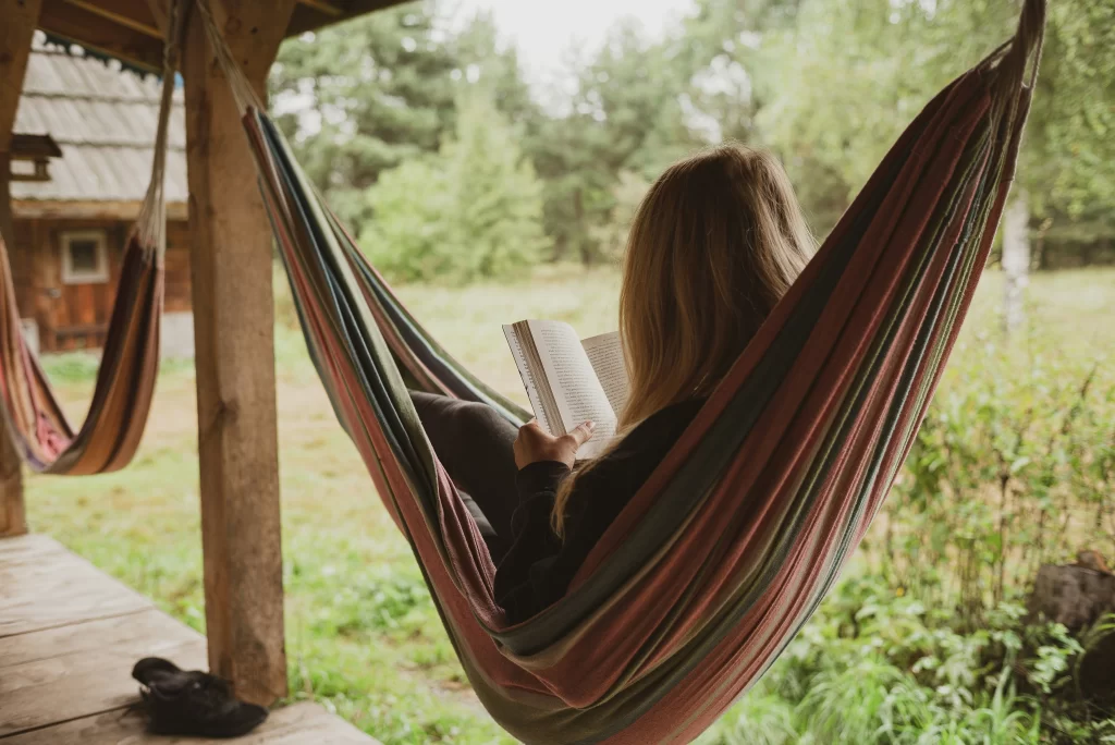 Personne vue de dos, allongée dans un hamac suspendu sous un porche en bois, lisant un livre. L’environnement est calme et naturel, avec de la verdure et des arbres en arrière-plan, évoquant un moment de détente et de lecture en plein air.