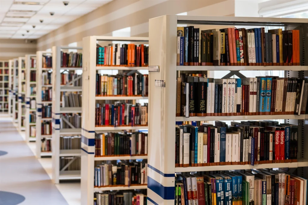 Une vue en perspective d'une allée de bibliothèque moderne montrant plusieurs rangées d'étagères blanches remplies de livres multicolores.