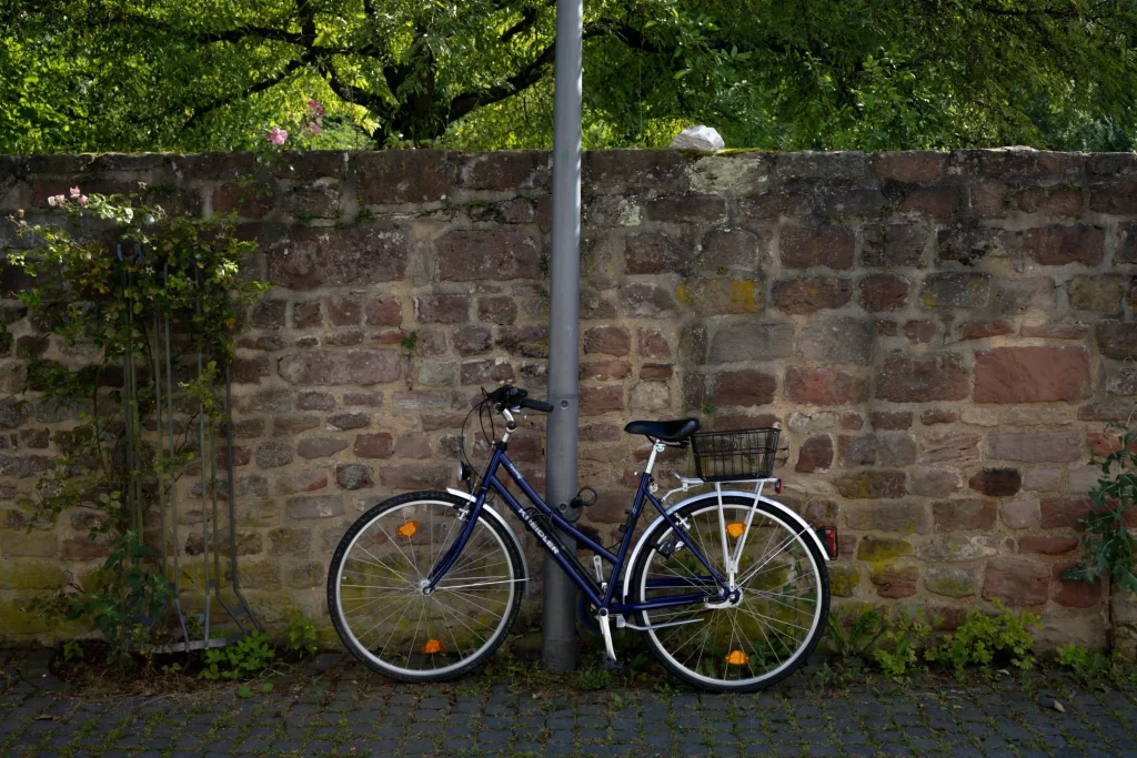 Un vélo de ville bleu foncé avec un panier arrière, stationné contre un poteau devant un vieux mur en pierre décoré de roses.