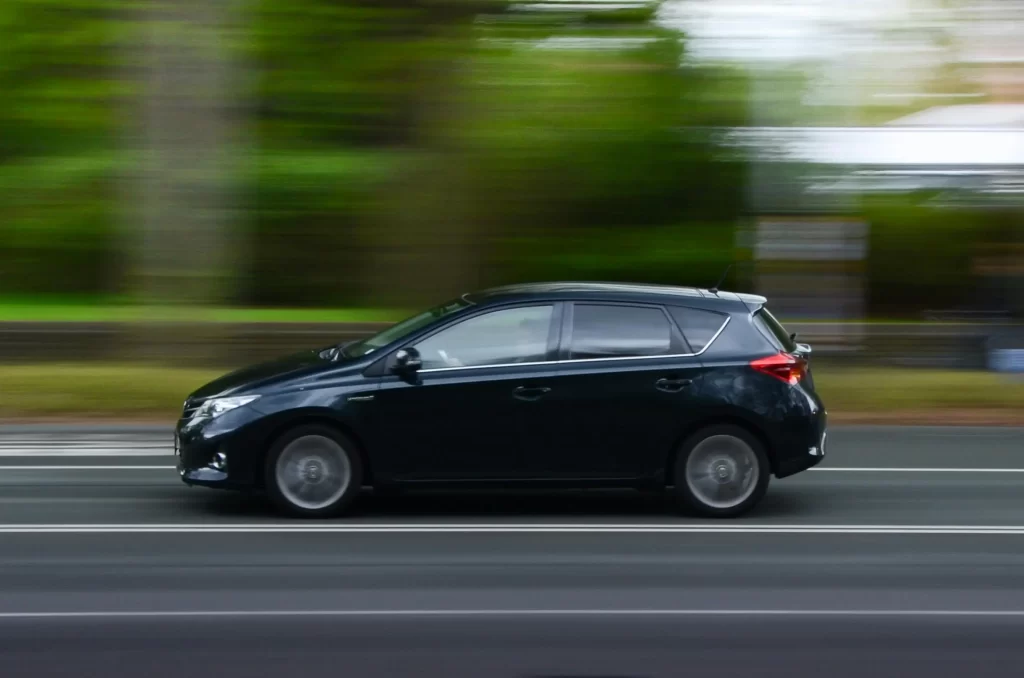 Vue de profil d'une voiture compacte noire roulant sur une route, avec un arrière-plan d'arbres verdoyants flouté par la vitesse.