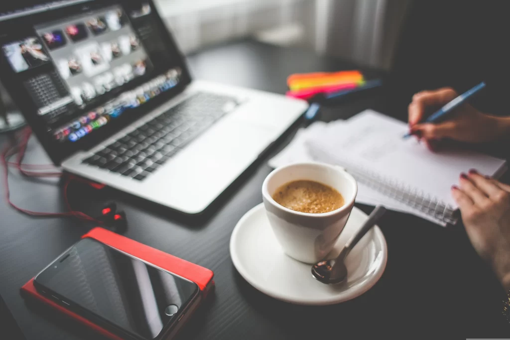 Une tasse de café au premier plan sur un bureau noir, avec un ordinateur portable ouvert et une personne prenant des notes en arrière-plan.