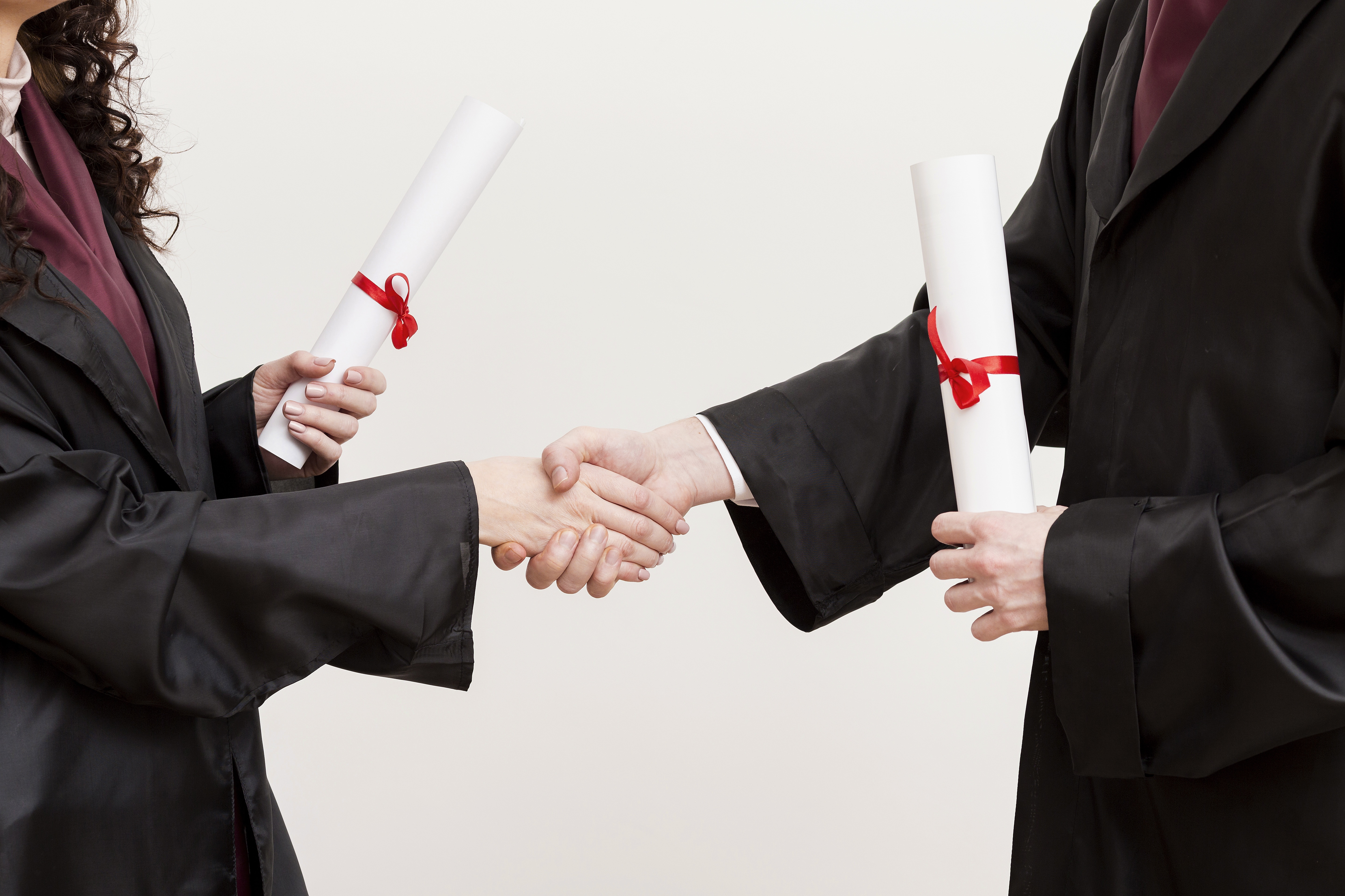 Deux personnes portant une toge de graduation se serrent la main en tenant chacune un diplôme roulé avec un ruban rouge.