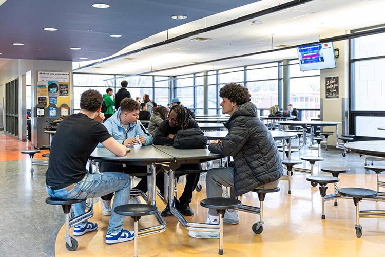 Quatre étudiants discutant autour d'une table ronde dans une cafétéria spacieuse et lumineuse. En arrière-plan, d'autres étudiants sont visibles près de grandes fenêtres.