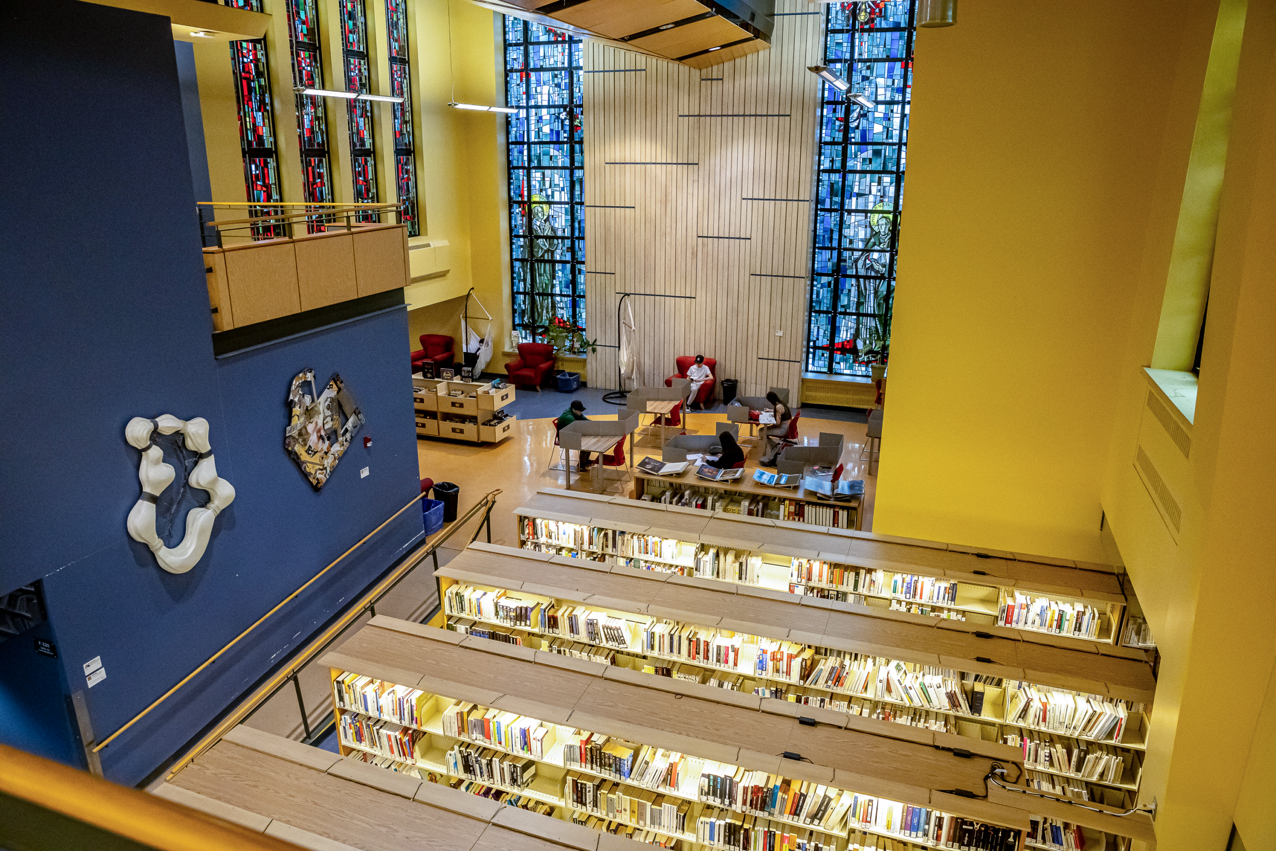 Une bibliothèque spacieuse avec de hauts plafonds et de grandes fenêtres en vitrail. On y voit des rangées de livres au premier plan, ainsi que des étudiants qui étudient à des tables et dans des fauteuils confortables à l'arrière-plan.