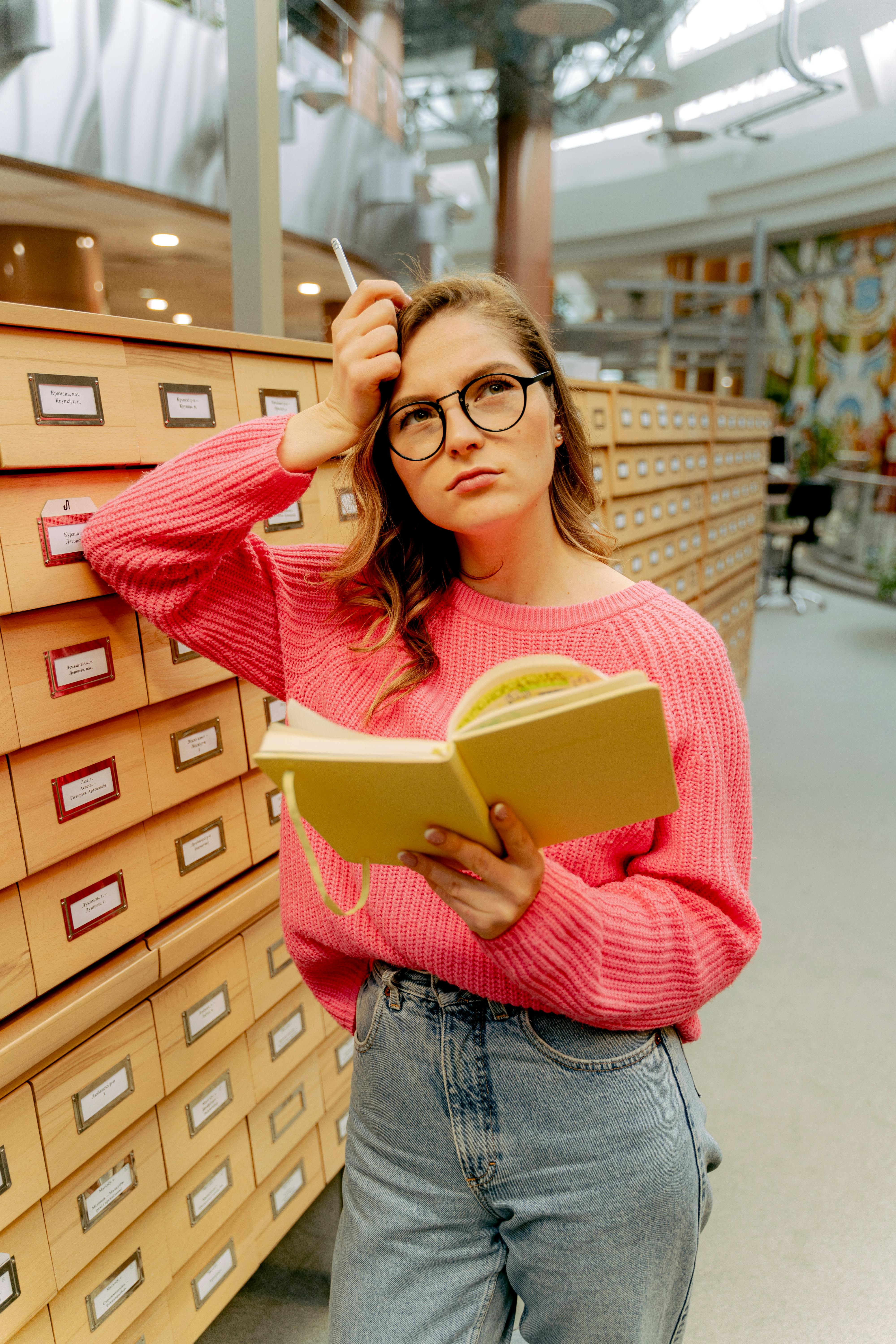 Jeune femme portant des lunettes et un pull rose, l'air pensive, consultant un carnet devant un meuble d'archives à tiroirs dans une bibliothèque ou une université.