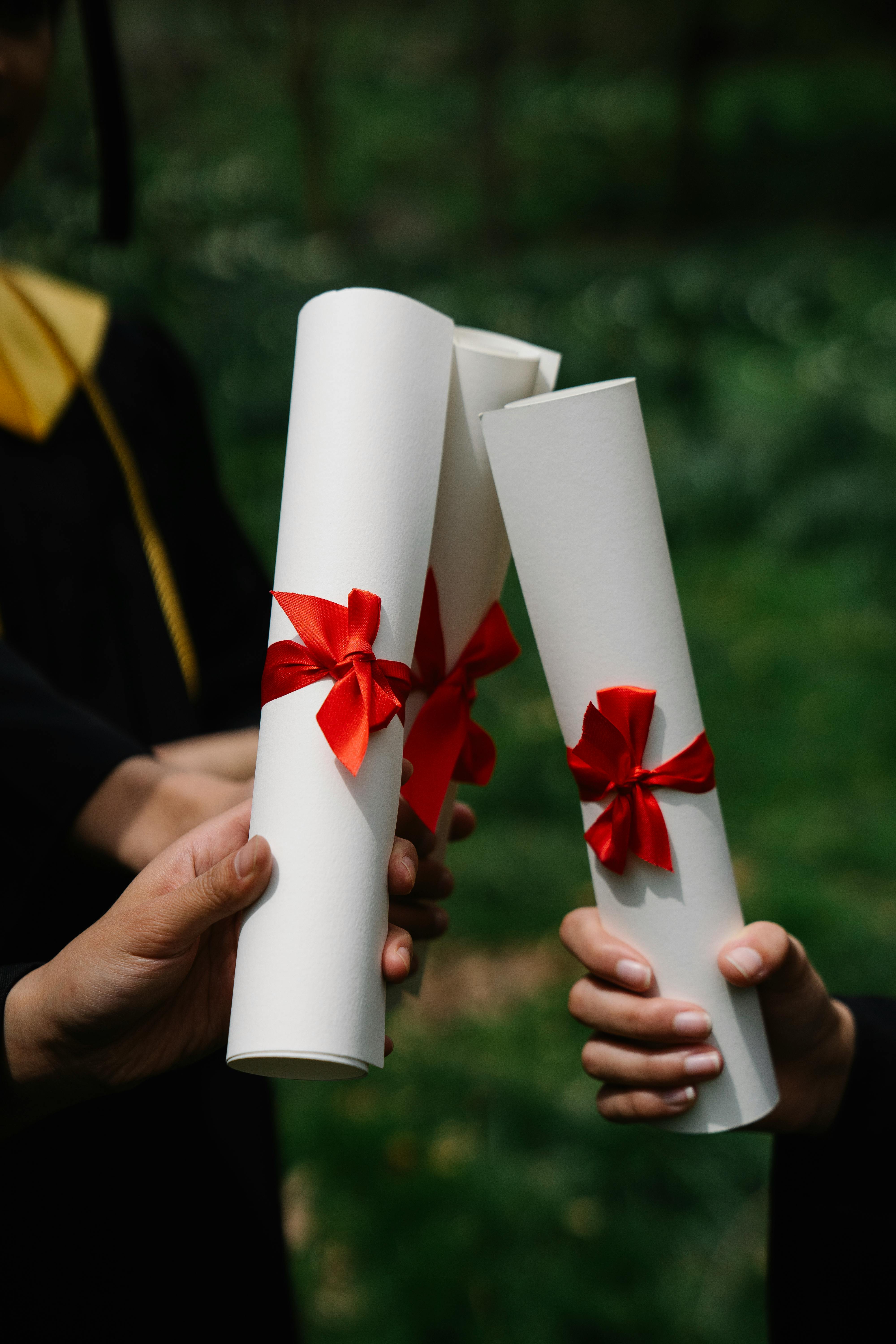 Gros plan de deux mains d'étudiants tenant fièrement trois diplômes roulés et attachés avec des rubans rouges. À l'arrière-plan flou, on aperçoit une robe de remise des diplômes noire et dorée et un jardin verdoyant.