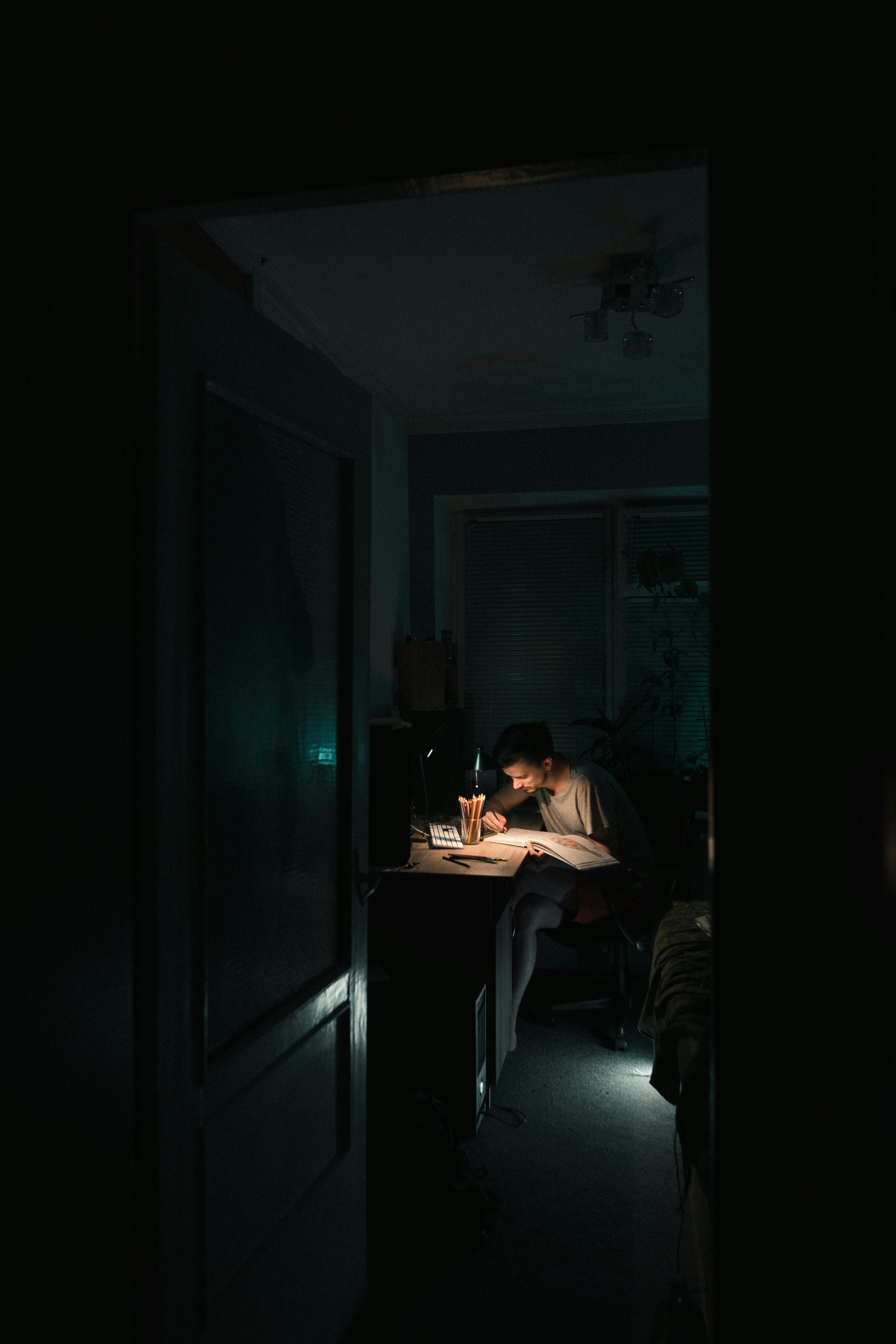 Vue à travers une porte d'un jeune homme étudiant tard le soir dans une chambre sombre, éclairé uniquement par sa lampe de bureau, symbolisant la concentration et le travail nocturne.